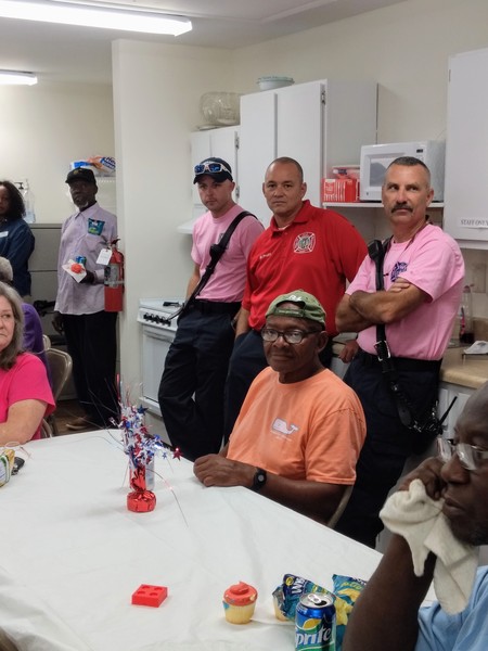 Attendees gathered around a table while Emergency Workers stand behind them, two wearing pink shirts and one wearing a red shirt
